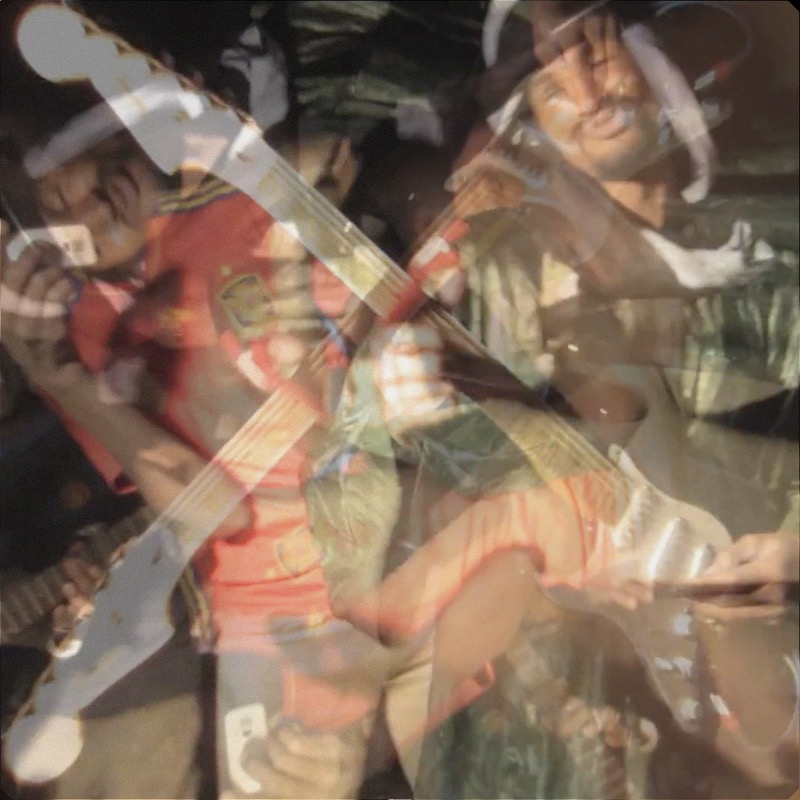 A warm square photograph of Mdou Moctar playing an electric guitar in formal Saharan clothing, surrounded by people dancing outdoors in the dark under harsh directional lighting. The veil combine creates a soft double-exposure effect — a translucent ghost of the same scene is laid over itself, blending the warm oranges, browns, and earth tones into a diffuse dreamlike doubling across the full image. There is no distinct distortion region or noise.