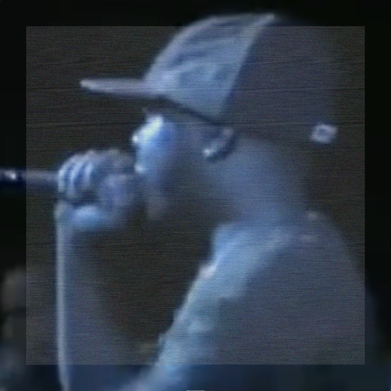 A lofi close-up of J Dilla holding a microphone to his mouth, looking left, wearing a ballcap in a live stage setting. The midpoint combine and angled traversal have cast the entire image in a uniform cool blue-gray wash — there is no distinct distortion region, just a smooth blue-toned rendering of the original low-resolution footage.