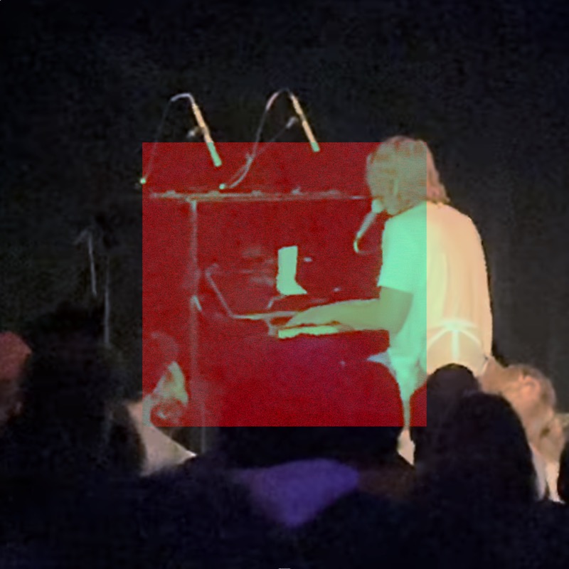 A medium shot of Cameron Winter playing an upright piano on a spotlit stage, facing to the left and away from the audience. He is wearing a white T-shirt, there are two directional microphones hovering above the piano like antennae, facing down. The back of the heads of many audience members are at the base of the image. A centered distortion square covers two thirds of the image increasing the presence of reds on the primarily blue and white image.