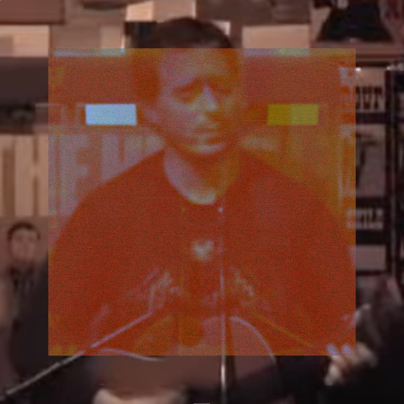 A medium shot of Jason Molina seated at a microphone, head bowed downward, in a record store — shelves of records and posters visible in the background. A large orange-red rectangle covers the central two-thirds of the image in warm distortion. Within the rectangle, two small isolated bright blocks appear — one pale blue and one yellow — like corrupted color artifacts against the orange wash. The outer edges retain the natural warm brown tones of the shop.